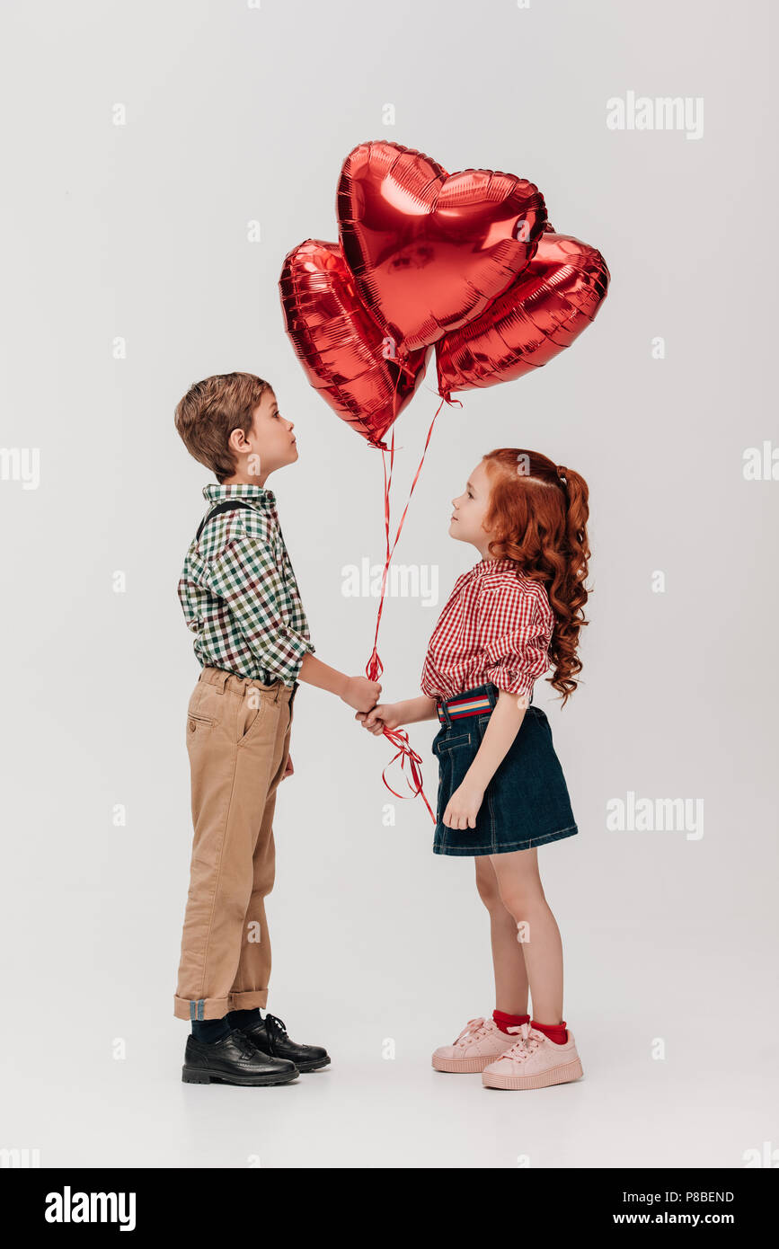 side view of beautiful little couple holding heart shaped balloons ...