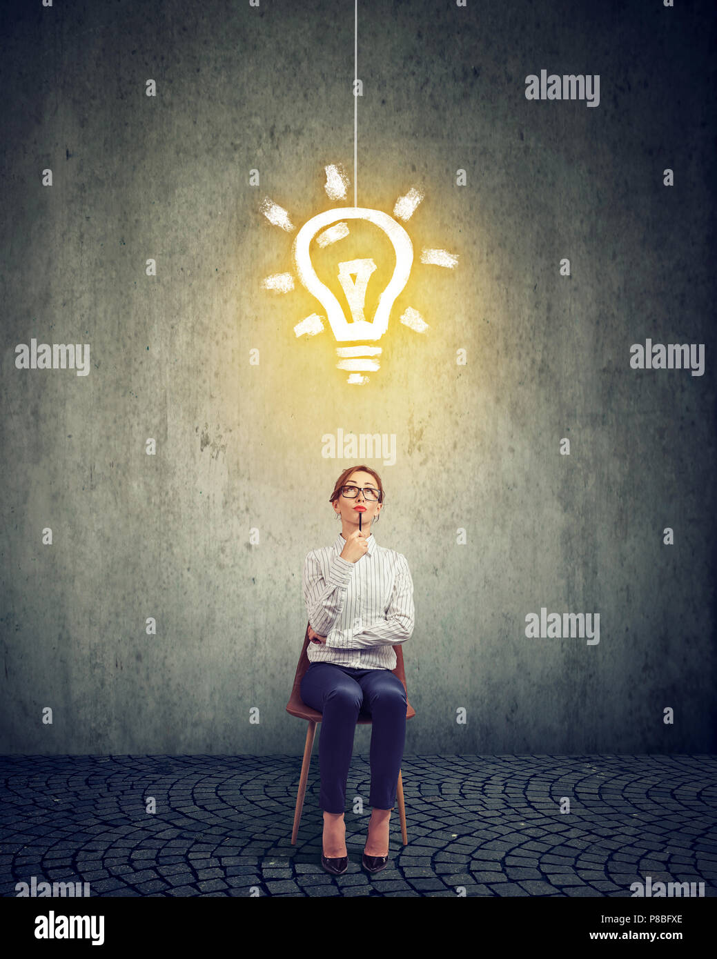 Stylish thinking businesswoman creating new strategy and looking up at bright light bulb while sitting on chair against gray background Stock Photo