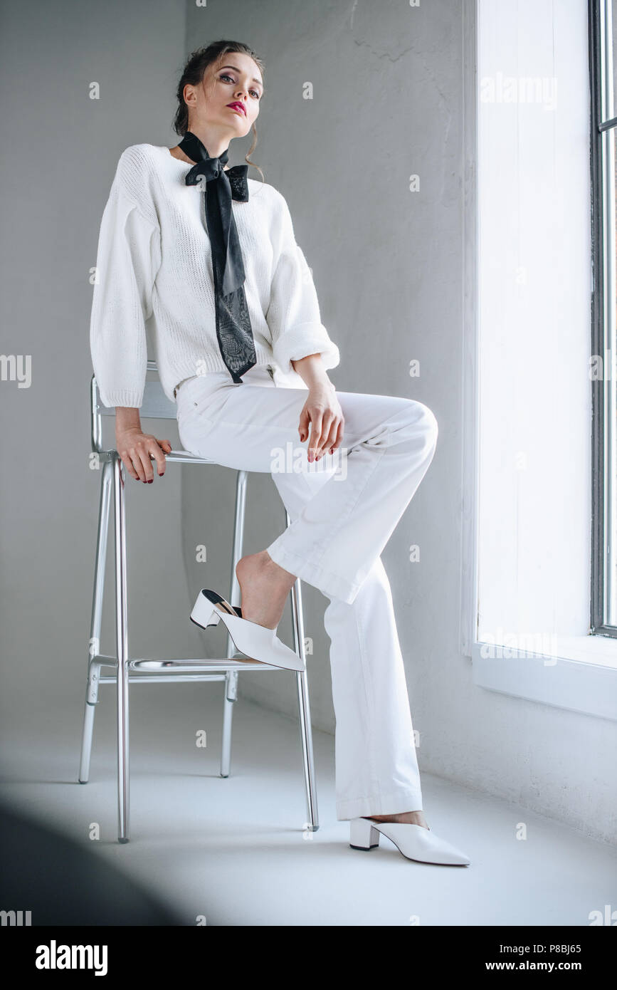 full length view of stylish young woman in white clothes with black bow on neck sitting on chair and looking at camera Stock Photo