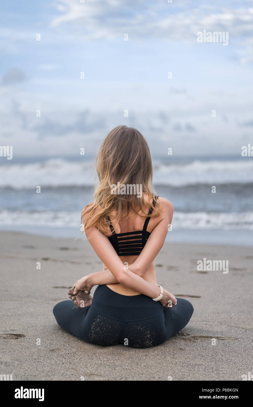 rear view of young woman practicing yoga in lotus pose with arms crossed behind back on seashore Stock Photo