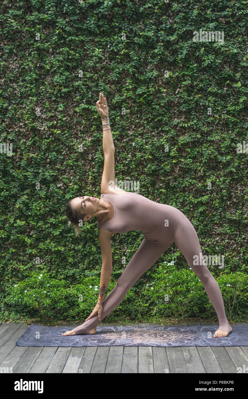 side view of beautiful young woman practicing yoga in Revolved Triangle pose (Parivrtta Trikonasana) in front of wall covered with green leaves Stock Photo