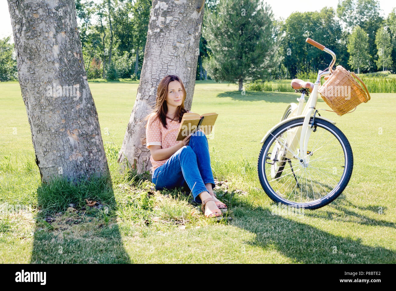 Beautiful carefree woman sitting under a tree with a book on a summer day Stock Photo