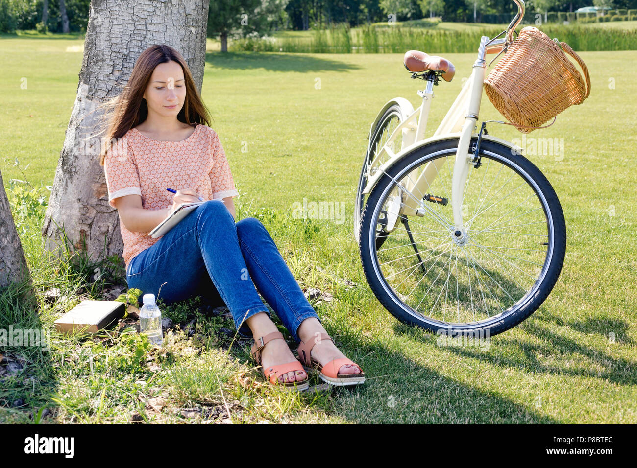 Young happy woman student writes in a notebook sitting under a tree on a summer day Stock Photo