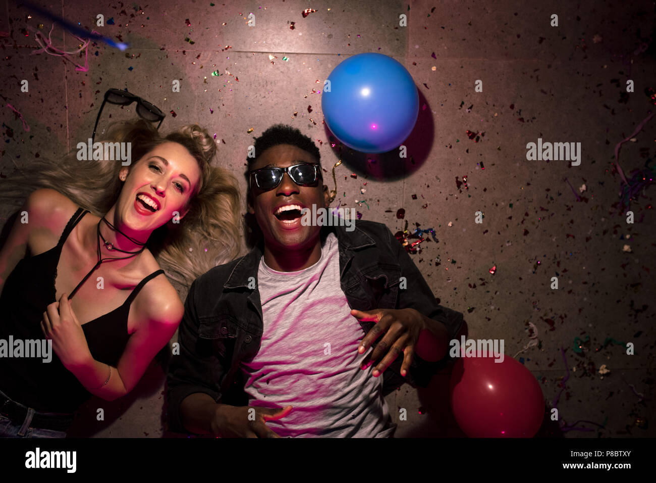 Top view of man and woman lying on floor and laughing at a house party. Tired friends lying on floor at the house party with balloons and confetti aro Stock Photo