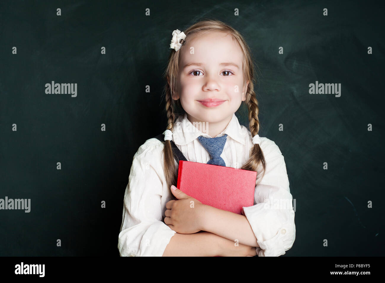 Cute little schoolgirl child with school book on green chalkboard background. Smiling pupil in classroom (child 5-6 years old) Stock Photo