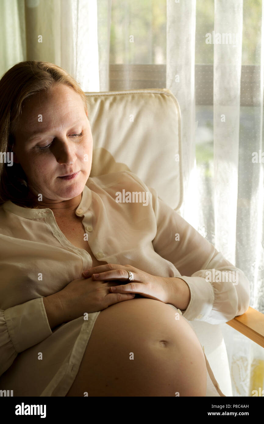 Blonde lady in mid-thirties sitting and caressing her pregnant belly. Stock Photo