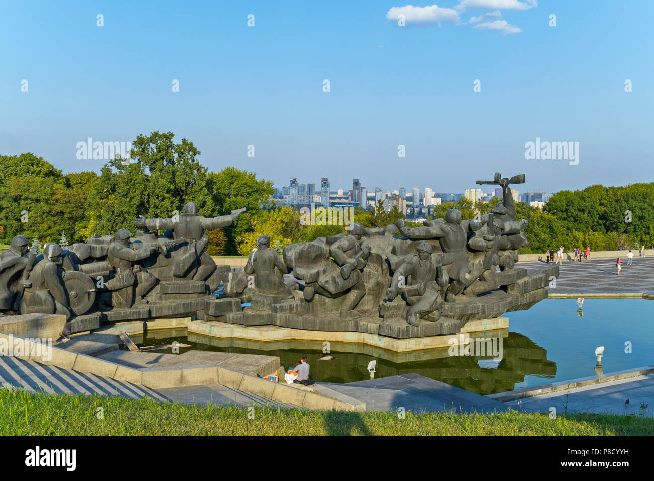 Memorial in the form of advancing soldiers made of stone. Standing in the park on the water against the backdrop of green trees and city quarters in t Stock Photo