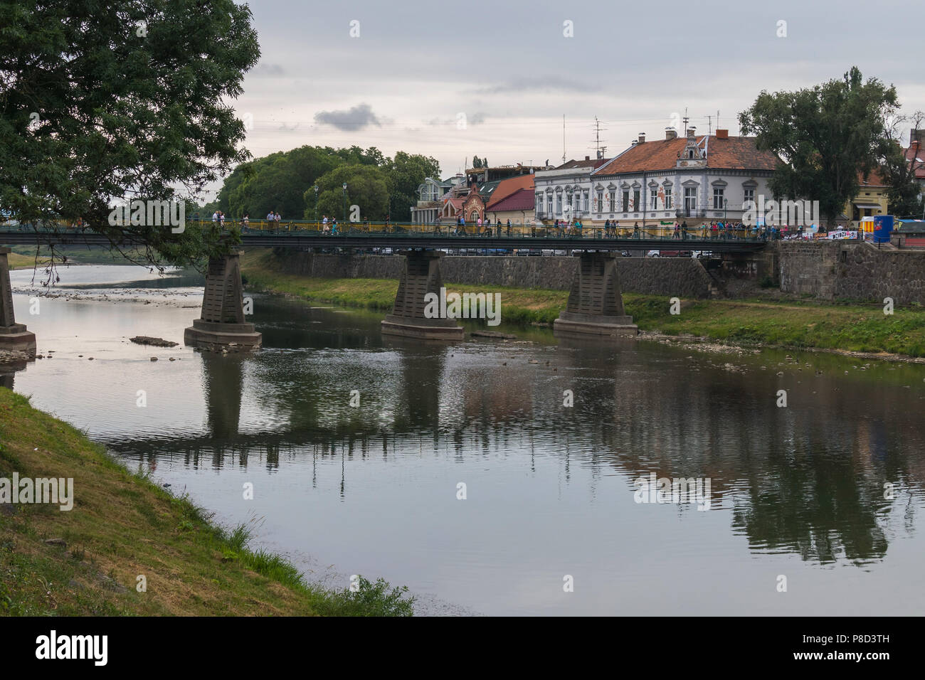 A large pedestrian bridge across the river with many people strolling along it and the embankment. Against the background of houses with ancient archi Stock Photo