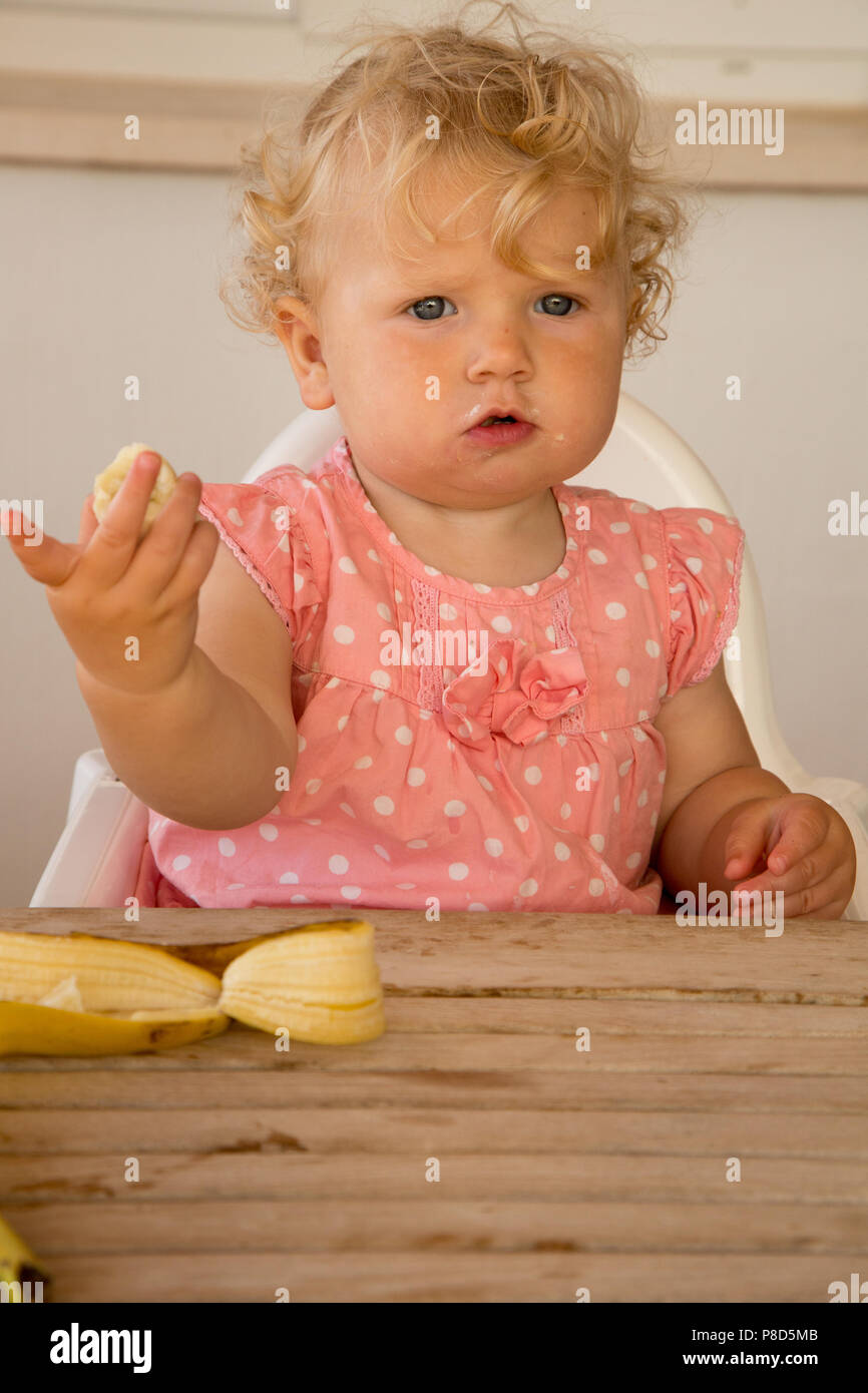 A one year old girl feeding herself a banana Stock Photo