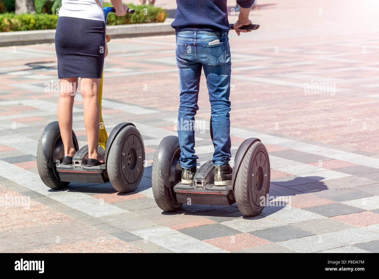 Couple in casual clothes riding modern gyro scooter hover board at city street on bright sunny day. Electric eco future urban transport gadget. Stock Photo