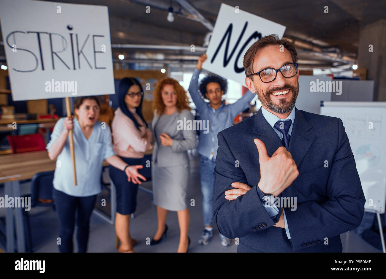 Laughing director showing thumb up, striking employees on backdrop. Modren loft office unsatisfied workers striking for their rights. Toned concept. Stock Photo