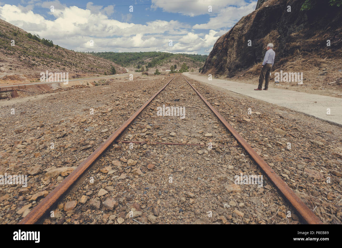Elderly man standing and facing away from the tracks of the train getting lost on the horizon in the Zaranda mines between the towns of Nerva and Riot Stock Photo