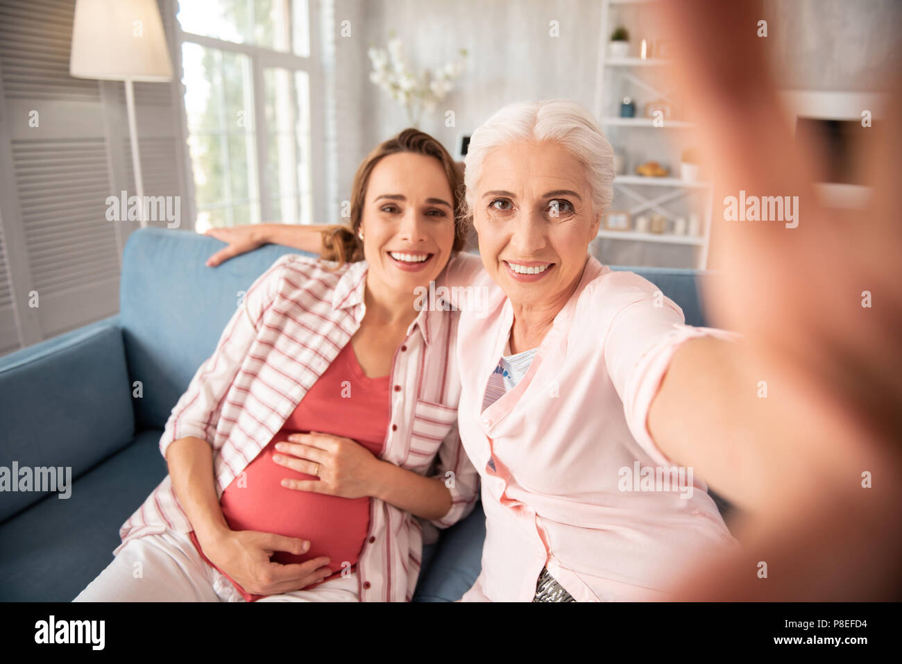 Pregnant woman wearing striped shirt sitting on sofa near her old friend Stock Photo