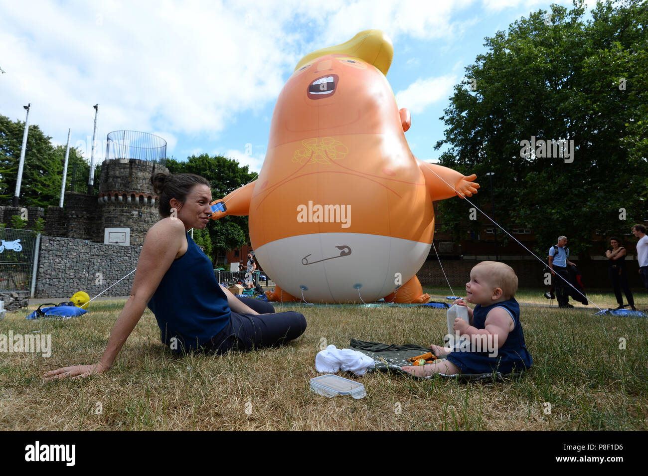 Nine-month-old Ivy Eakin and her mother Ruth sit in front of the Trump Baby Blimp as is inflated for a practice test, at Bingfield Park in north London. Stock Photo