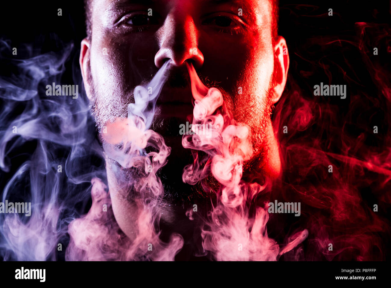 A young man exhales a cloud of colored smoke of red and pink color on a  black isolated. Harm to smoking on the example of a young mugger Stock  Photo