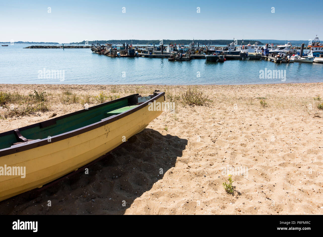 An empty rowning boat on the sand beside Poole Harbour with fishing vessels moored in the background, Poole, Dorset, United Kingdom Stock Photo