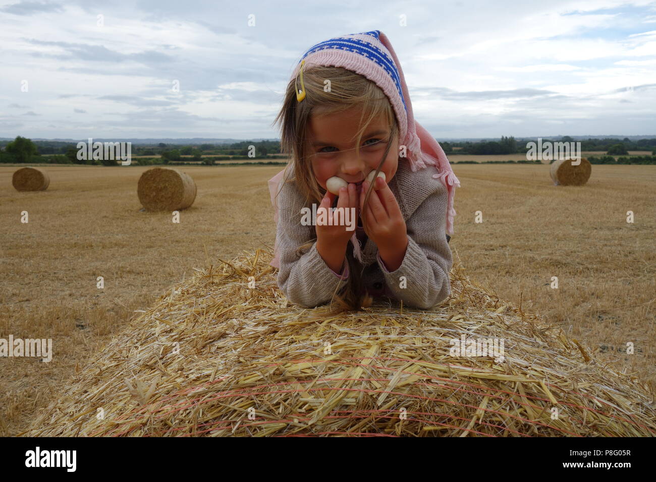 Beautiful girl in pink headscarf on straw bale in summer field at harvest time Stock Photo