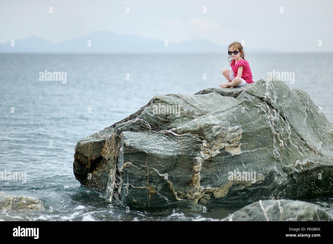 Adorable little girl sitting on a rock by a sea Stock Photo - Alamy
