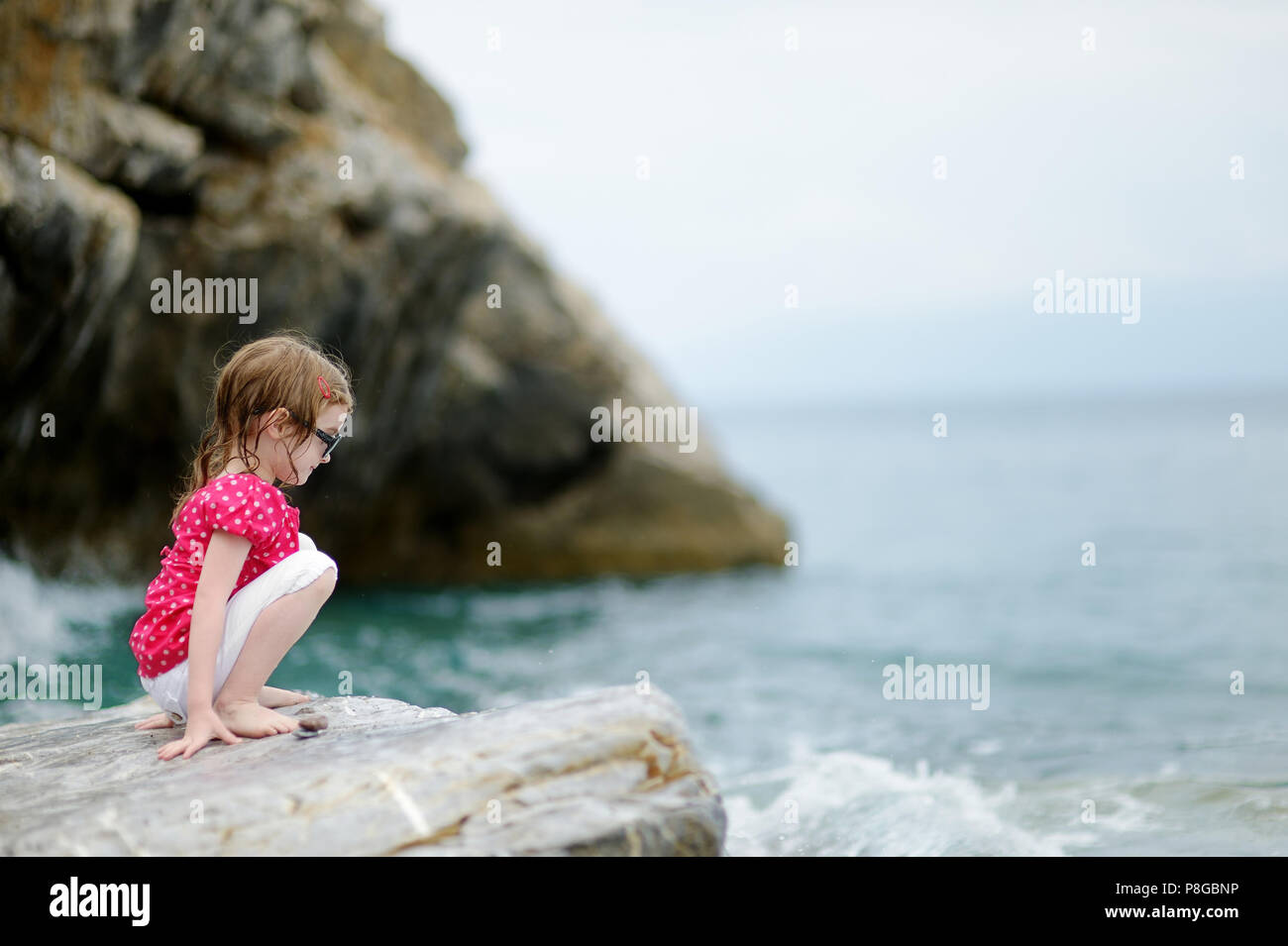 Adorable little girl sitting on a rock by a sea Stock Photo - Alamy