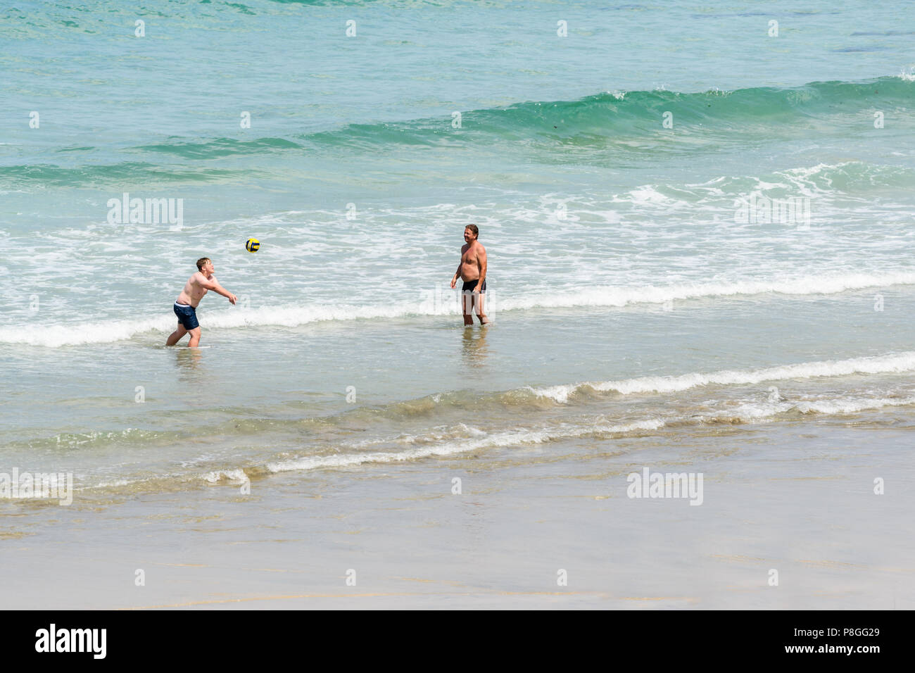 Two men play volleyball on the shore of the Atlantic ocean as its waves hit the sandy beach at the village of St Agnes, Cornwall, England. Stock Photo