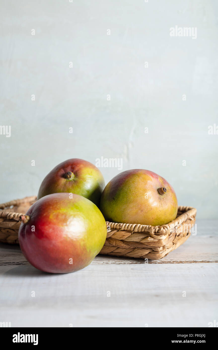 Three ripe mangoes in a wicker basket on a whitewash wood table with bright, natural light. Stock Photo