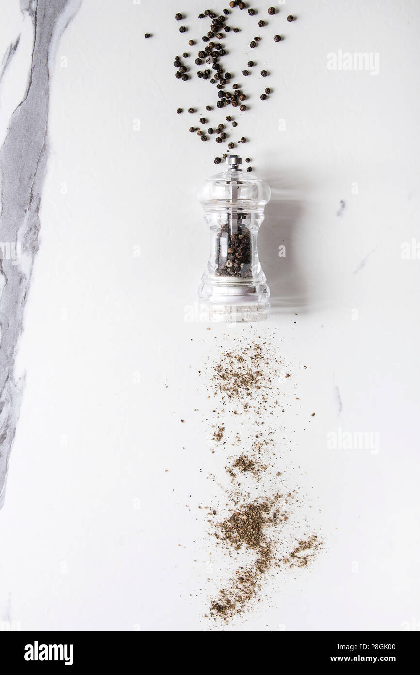 Black peppers peppercorns and ground powder from transparent pepper mill over white marble