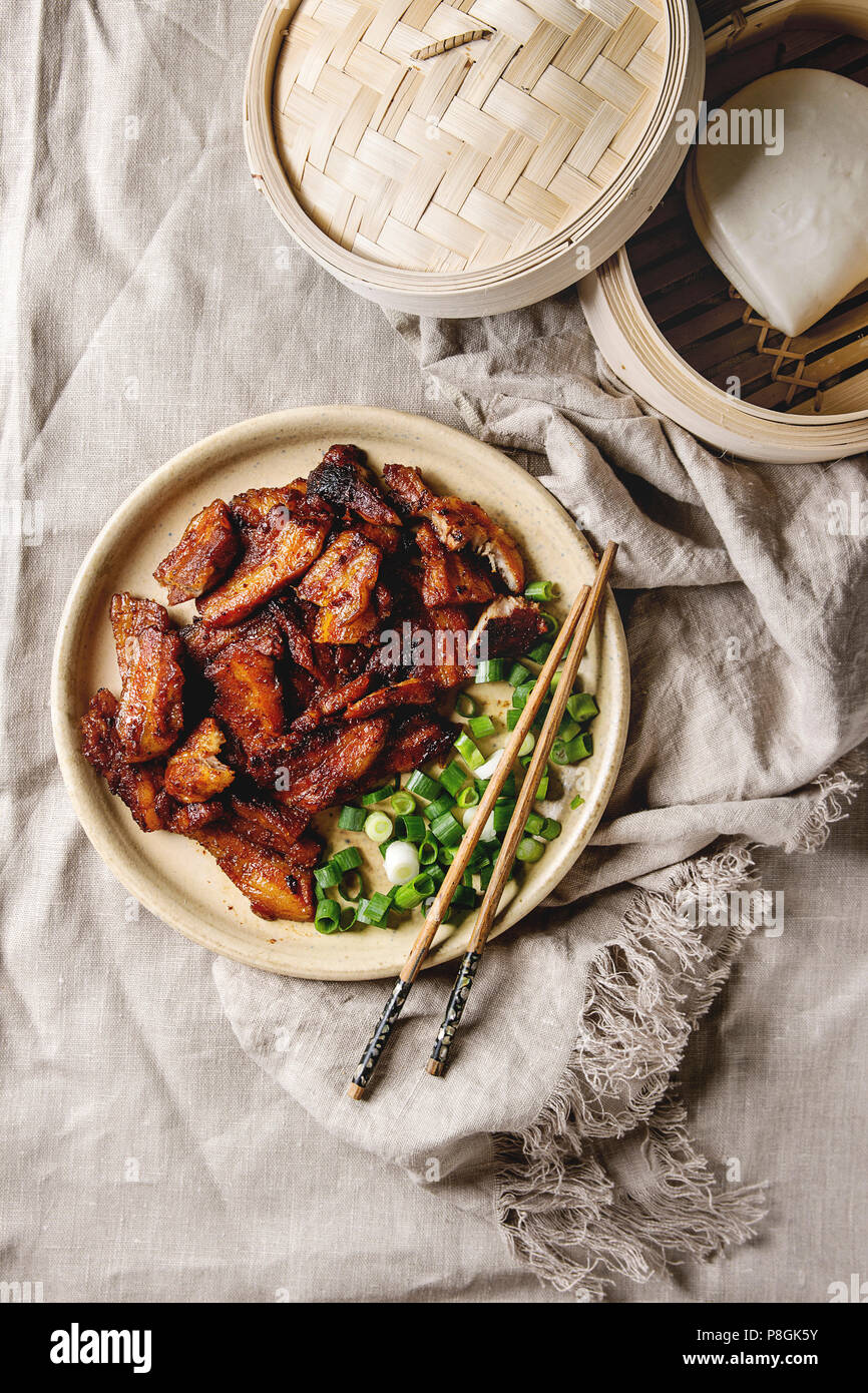 Chinese traditional dish Cantonese BBQ Pork Belly with spring onion served in ceramic plate with chopsticks, bamboo steamer and gua bao bus over linen Stock Photo