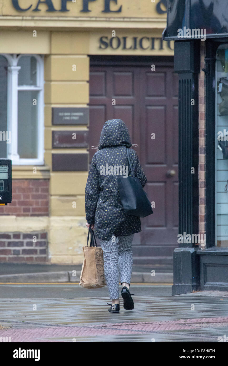 North Wales, 12th July 2018, UK Weather:  An overcast and cooler start to the day for some coupled with the first rain for many weeks with some showers being heavy and thundery, which may cause flooding. A woman walking to work in the rain wearing a rain coat with hood up in the market town of Mold, Flintshire, Wales, UK Stock Photo