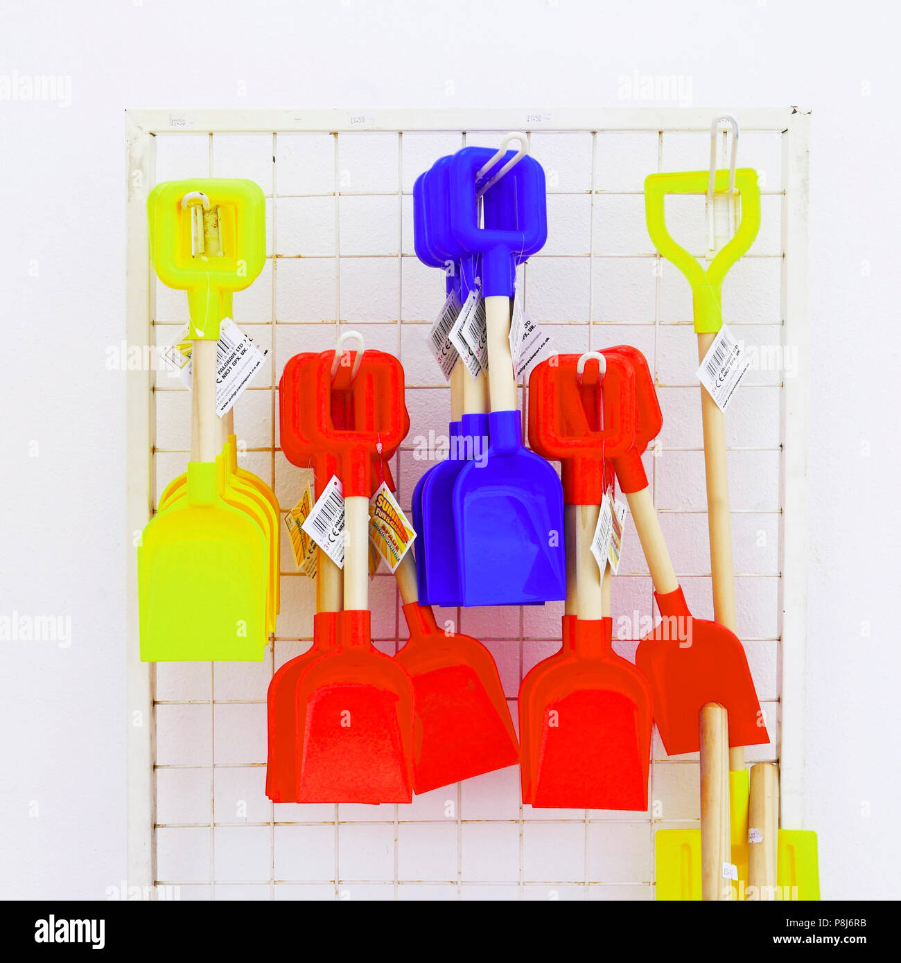 Red blue and yellow wood handled sand spades hanging on metal rack against white wall Stock Photo