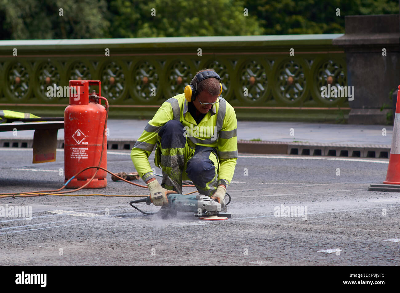 A road maintenance worker using an angle grinder power tool to finish off the road surface repairs. Worker is wearing ear defenders and high-vis top. Stock Photo