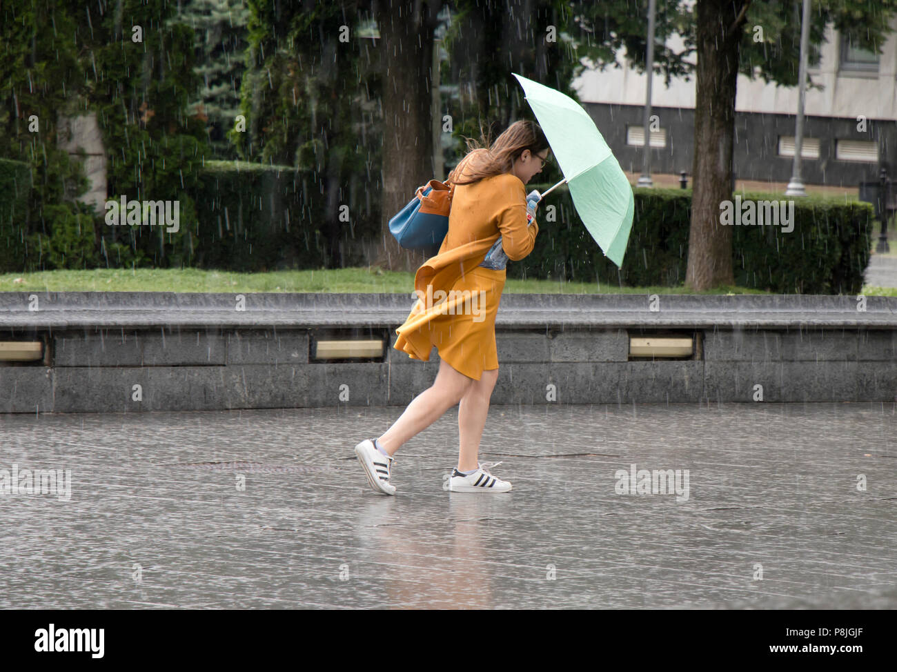 Belgrade, Serbia - June 14, 2018: One young woman running under umbrella in the sudden  heavy spring windy rain in the city park , holding a bottle of Stock Photo