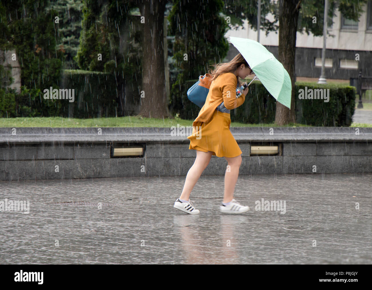 Belgrade, Serbia - June 14, 2018: One young woman running under umbrella in the sudden  heavy and windy  spring rain in the city park , holding a bott Stock Photo