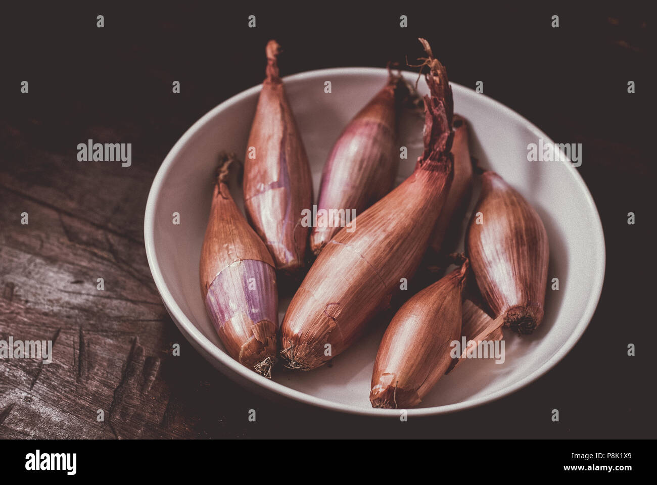 A moody Still life food image of Organic Shallots in a white bowl against a dark wooden background in  an Instagram style. Stock Photo