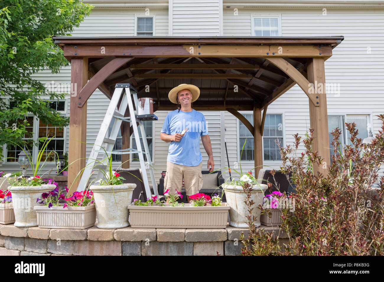 Pleased satisfied man staining a new gazebo installed on his rear patio in front of his house standing smiling at the camera Stock Photo