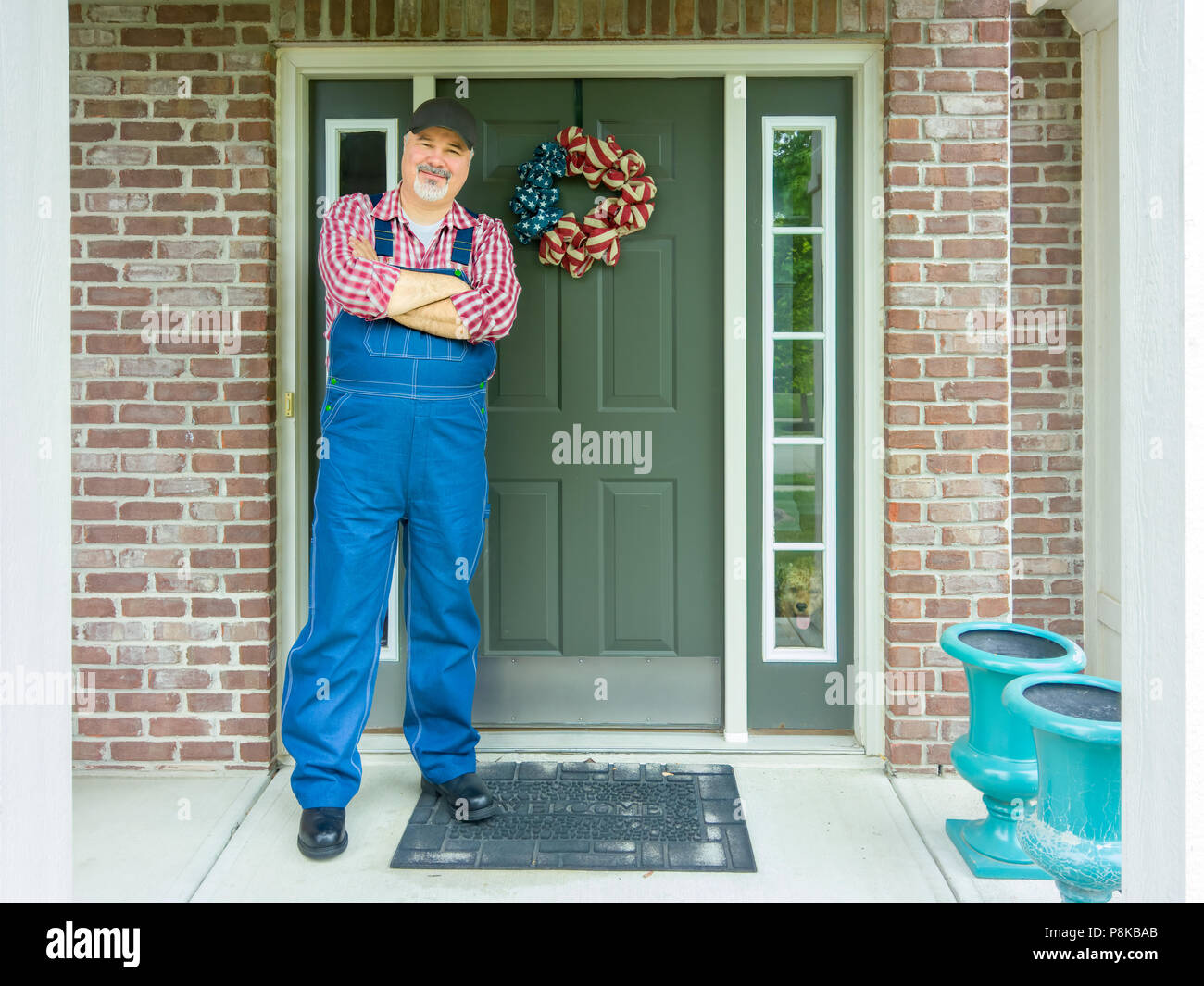 Patriotic farmer celebrating 4th July Independence Day standing with folded arms in front of the door of his farmhouse decorated with a wreath in the  Stock Photo