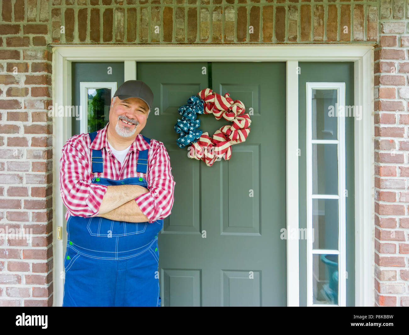 Happy friendly farmer wearing patriotic colors celebrating 4th July Independence Day in America standing in front of his door decorated with a wreath  Stock Photo