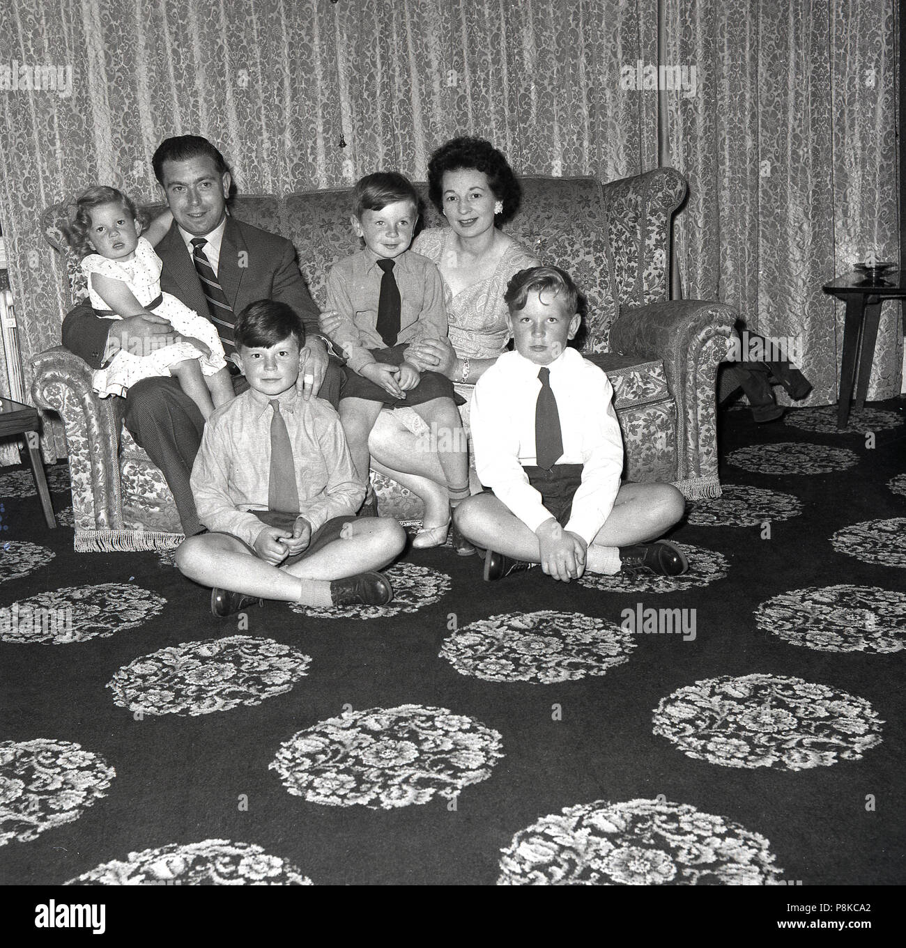 1950s, famliy at home, mother and father with their three boys and young infant sitting togetther on the sofa in their living room which has a distinctive patterned carpet. Stock Photo
