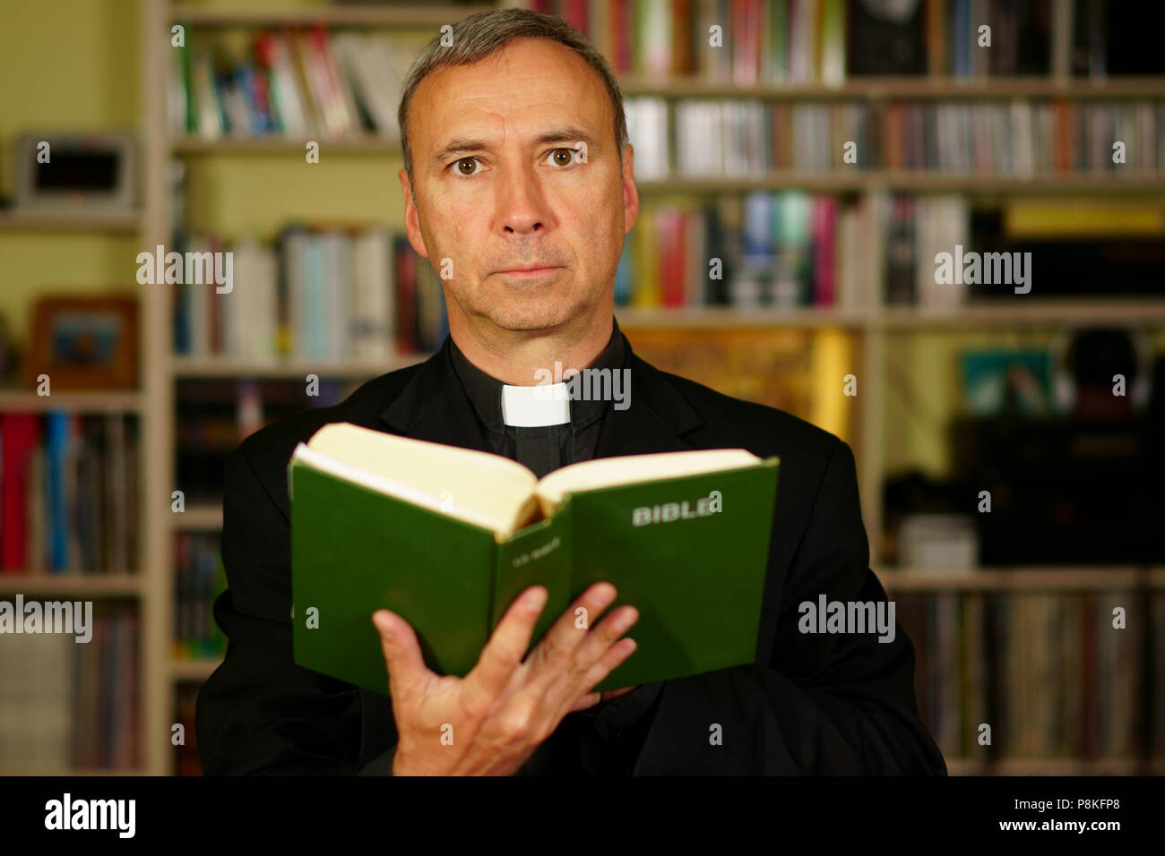 A good looking serious catholic priest is studying, reading the Bible, into his library. He looks at us. Stock Photo