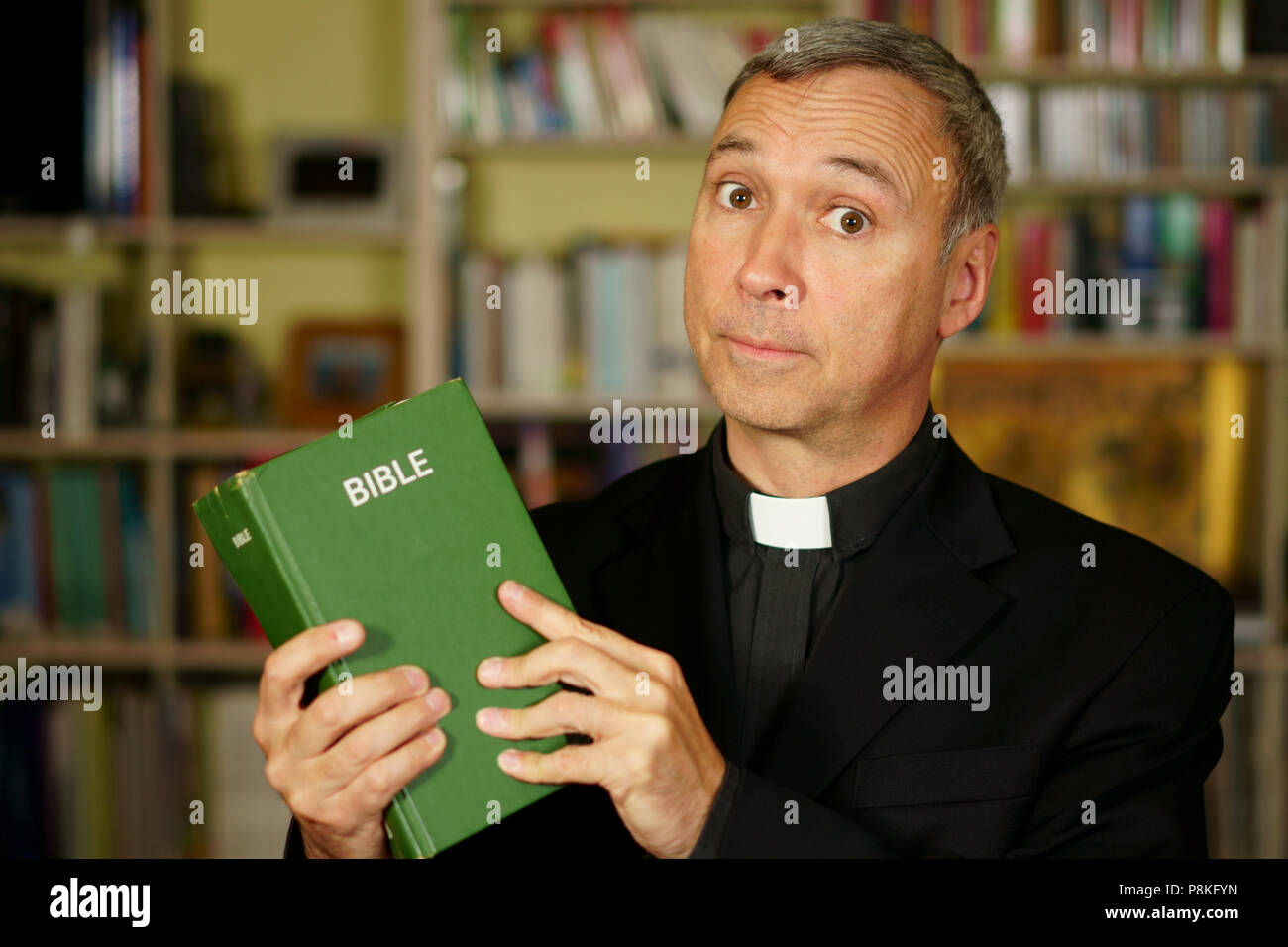 A good looking serious catholic priest is studying, reading the Bible, into his library. He looks at us with disapproval. Stock Photo