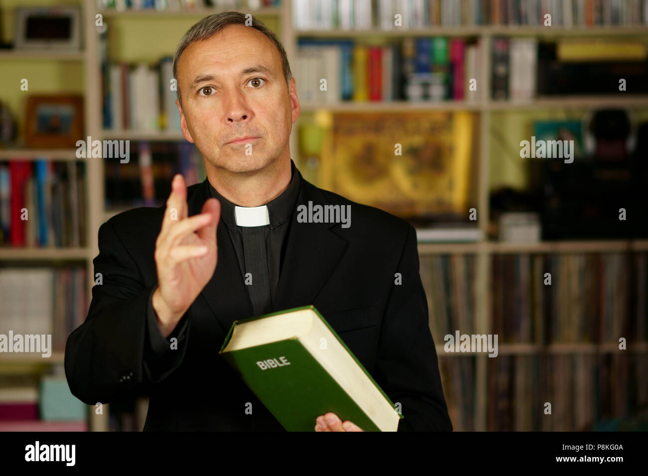 A good looking catholic priest is giving the blessing into his library. He seriously looks at us with  interest, disapproval, judgement, pensiveness a Stock Photo