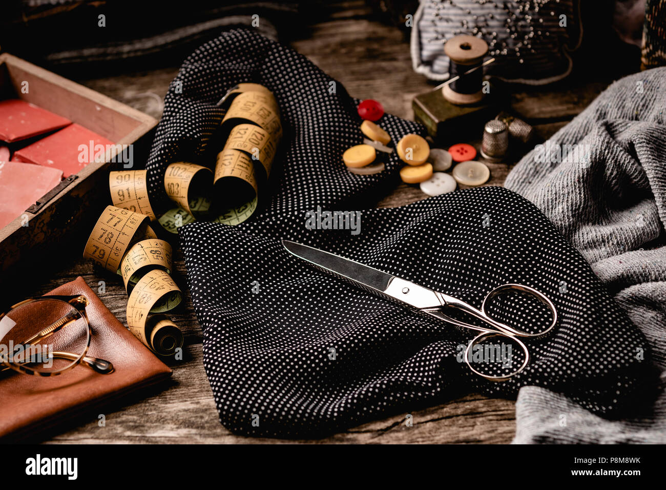 Composition of black polka-dotted fabric with scissors, sewing spools, tape measure and colorful buttons with glasses placed on rough wooden table Stock Photo