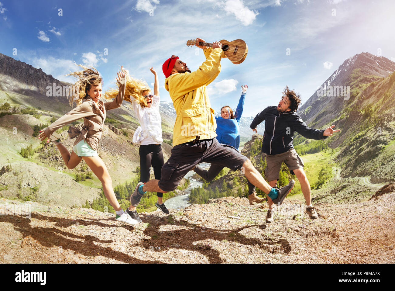 Group of happy friends with ukulele guitar is having fun and jumps against mountain during trekking. Music fun concept Stock Photo