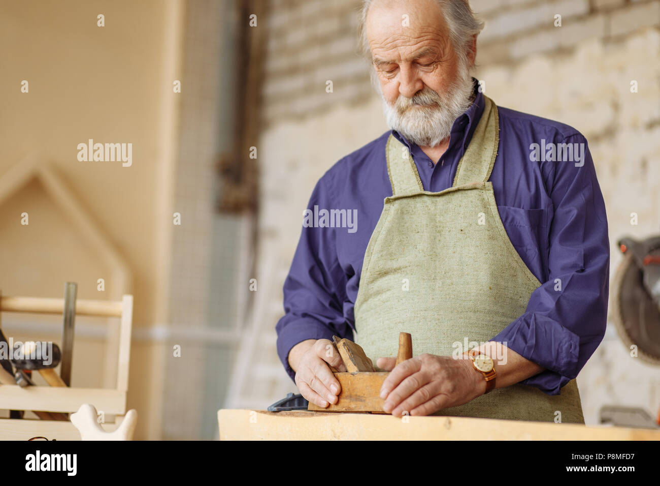 old experienced carpenter working with wooden planer on plank in workshop. Stock Photo