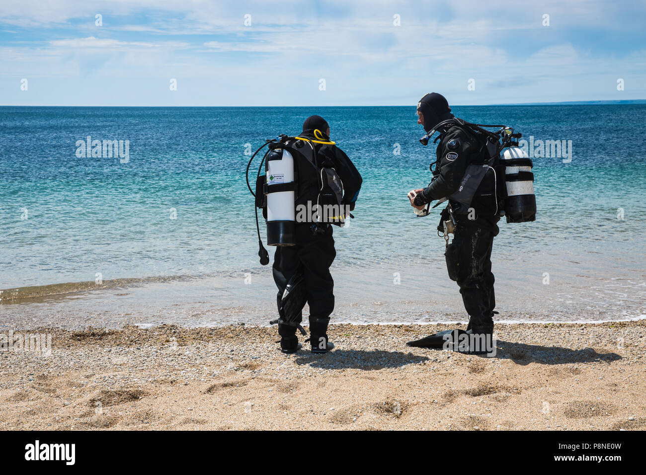 Two deep sea divers getting ready to dive by performing safety checks on their equipment together on a beach with the ocean behind Stock Photo