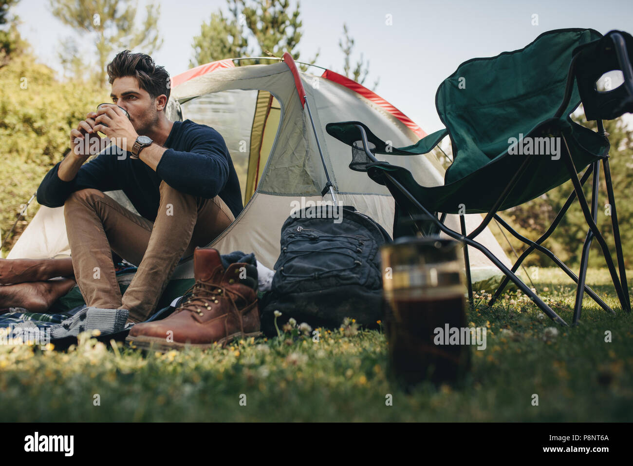 Young man sitting in his tent at campsite and drinking coffee. Male camper relaxing in a tent having a cup of coffee. Stock Photo