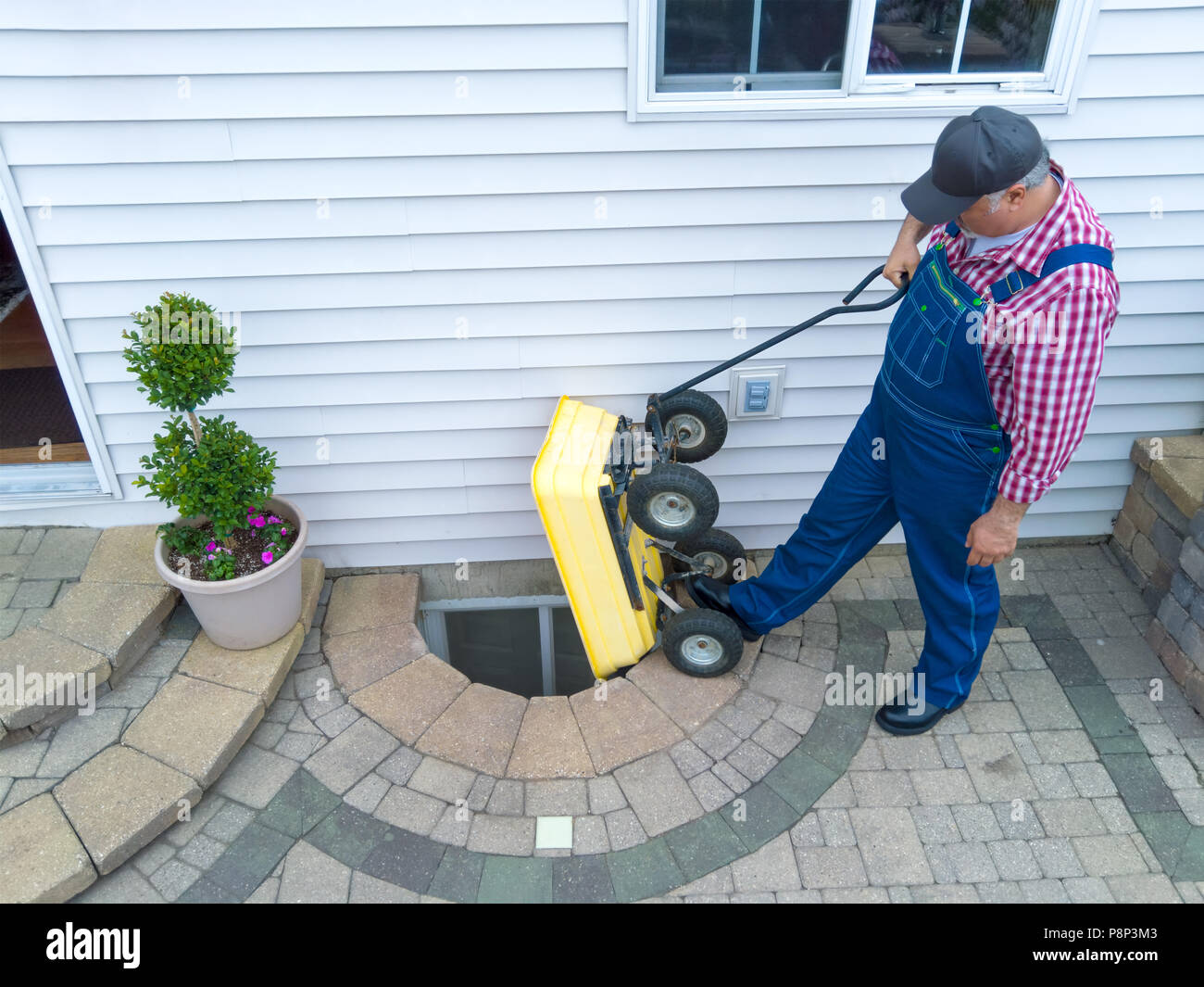 Man performing maintenance on a neglected egress window in the basement of his house emptying new pebbles from a wheelbarrow Stock Photo