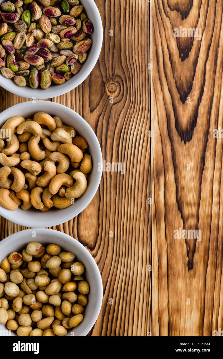 Three small white generic of assorted nuts with cashews, pistachios and peanuts on a rustic wood table with copyspace arranged as a side vertical bord Stock Photo
