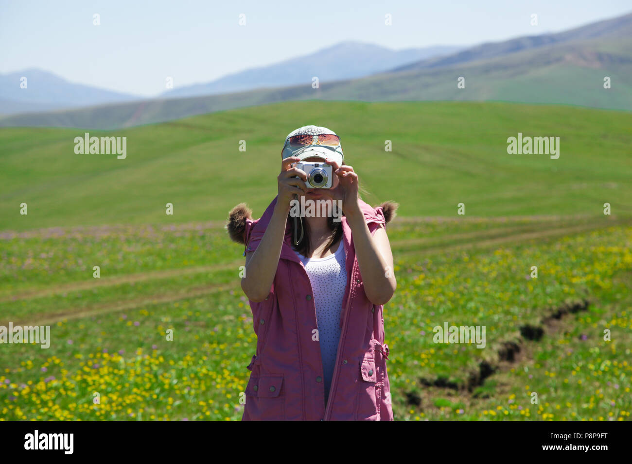 A woman in a pink waistcoat and a turquoise baseball cap photographs mountains Stock Photo