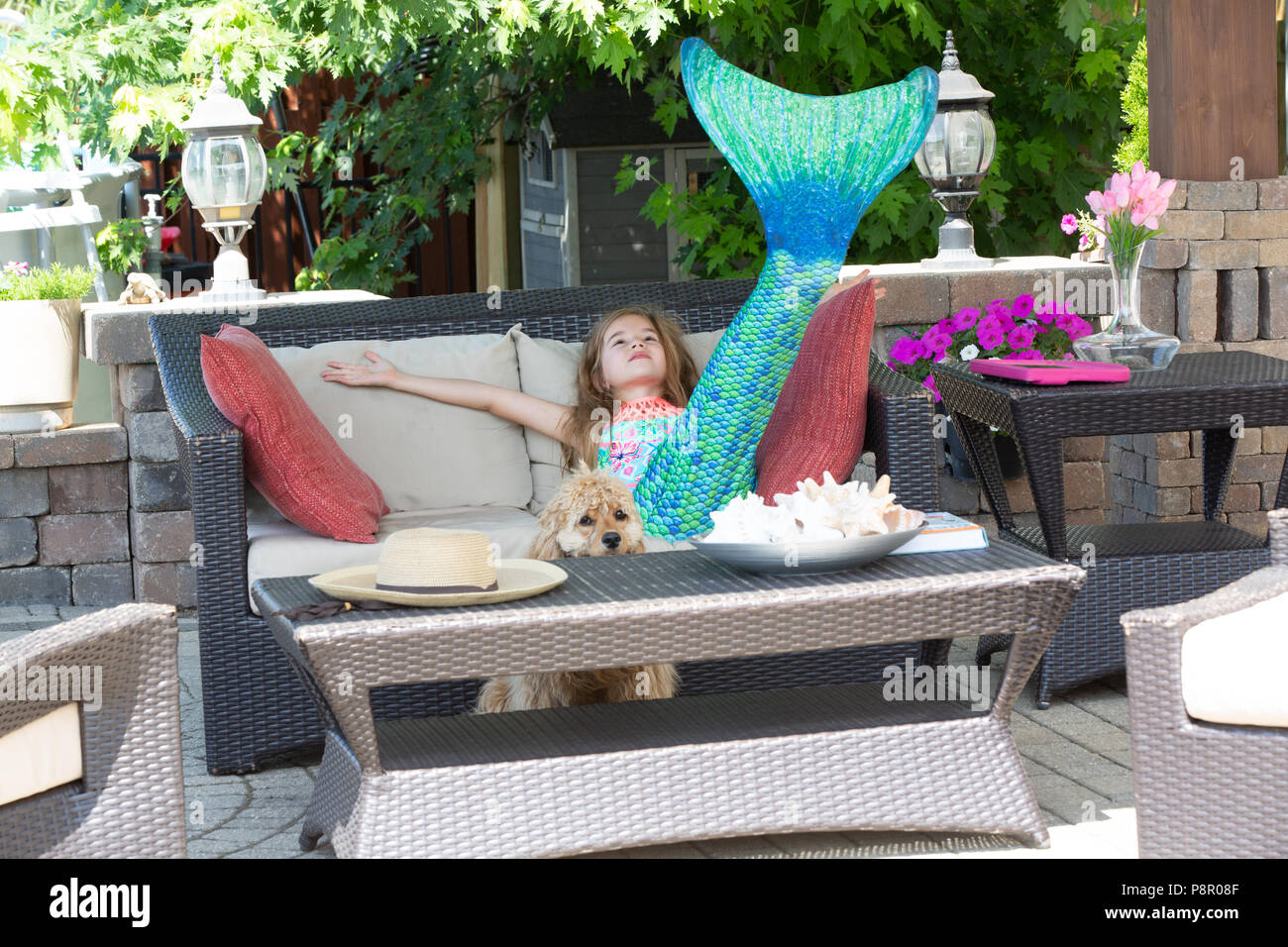 Young girl playing in a colorful mermaid outfit with a blue and green tail relaxing on a couch on the patio with her cocker spaniel puppy Stock Photo