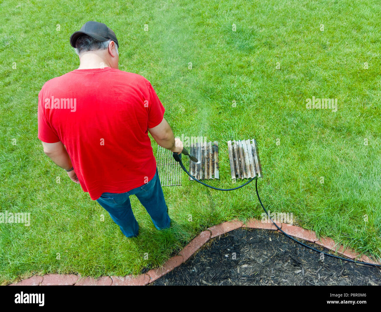 Man spraying a dirty barbecue grill with a pressure hose cleaning the fatty residue with a powerful jet of water on his lawn at home Stock Photo